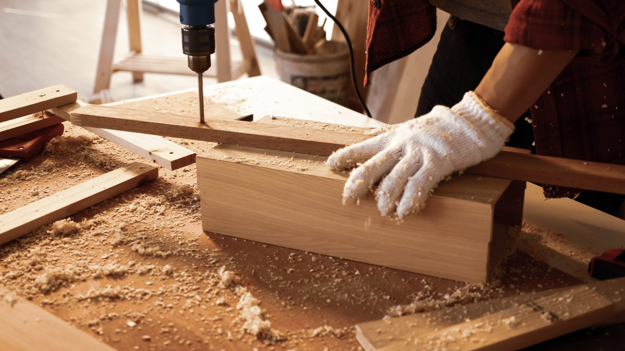 Person wearing gloves drills a hole in a wooden plank, surrounded by wood shavings on a workbench in a woodworking shop