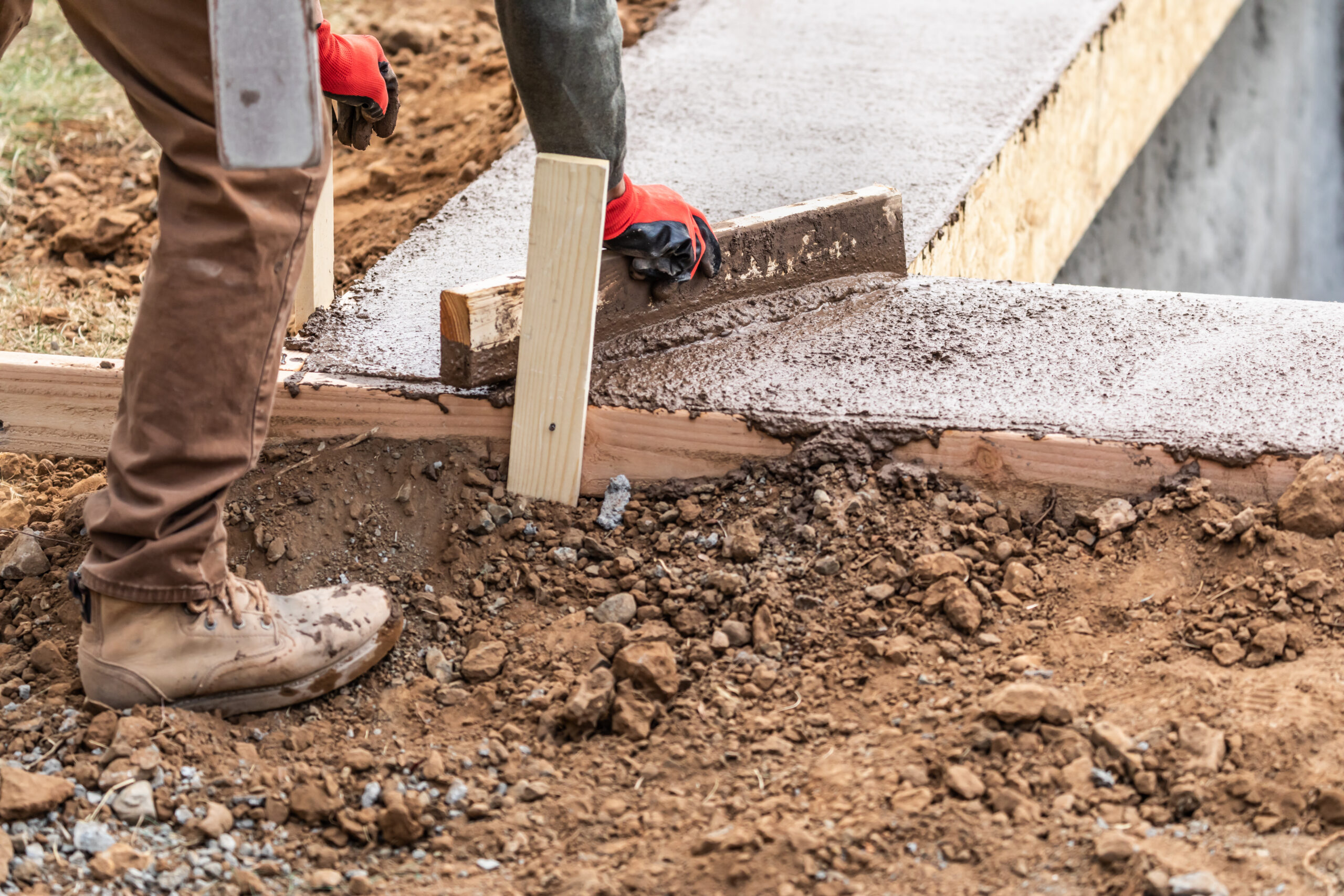 Construction worker's gloved hands and boots smoothing wet concrete along a wooden form.