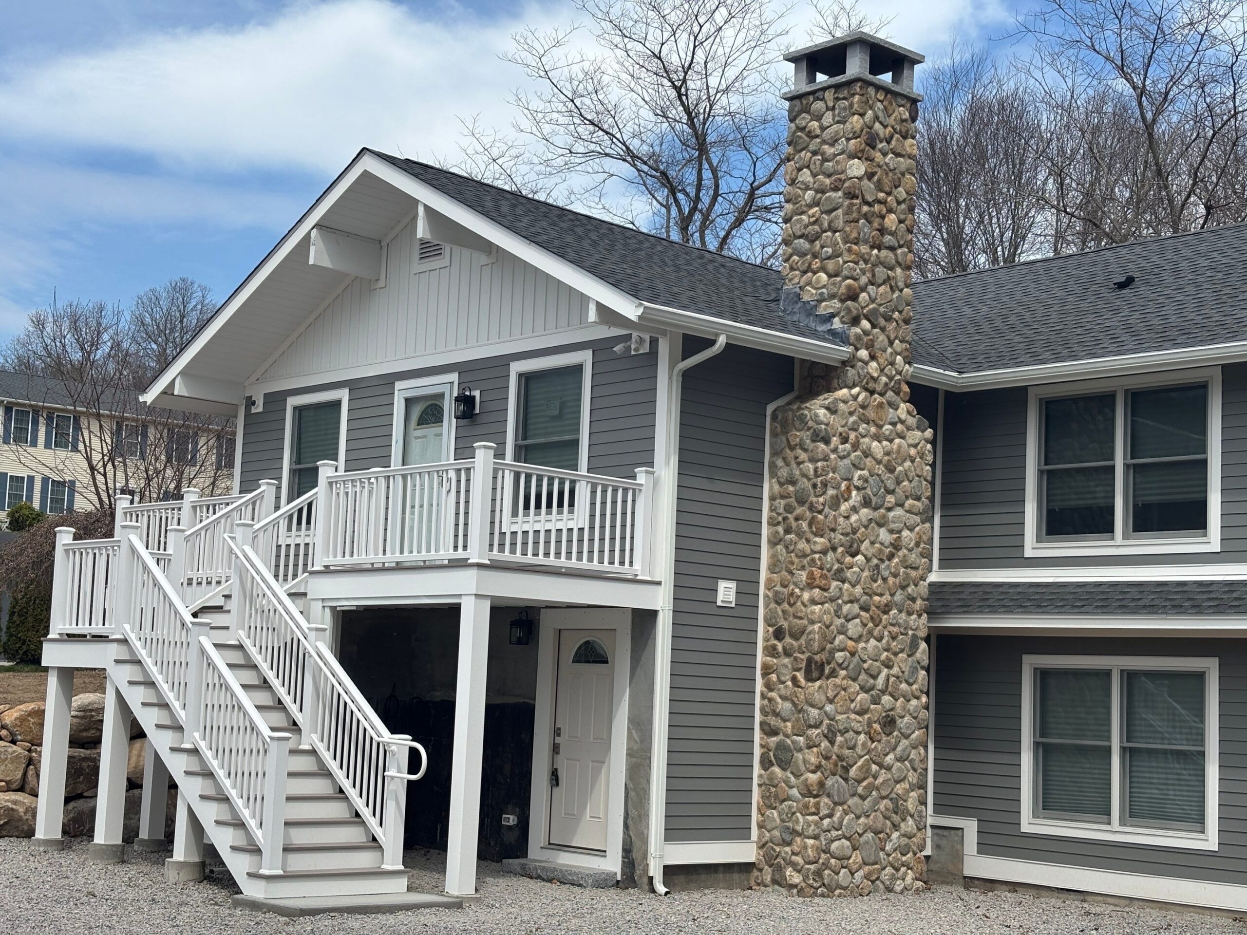 Front exterior of a gray house with a tall stone chimney, white railing and stairs, and a peaked roof under a blue sky.