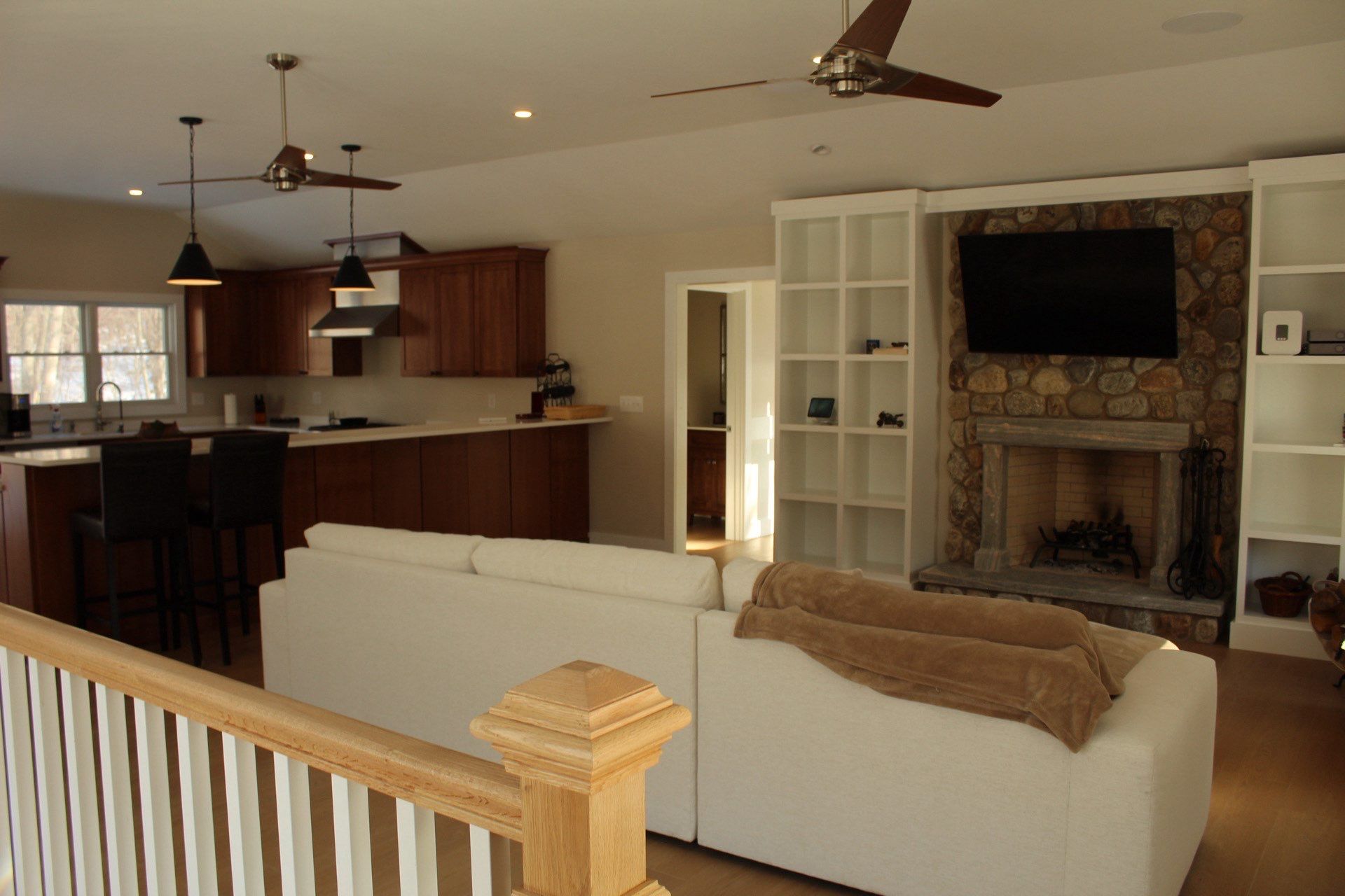Open-concept living area with white sectional sofa facing a stone fireplace and mounted TV, adjacent to a kitchen with dark wood cabinets and island.