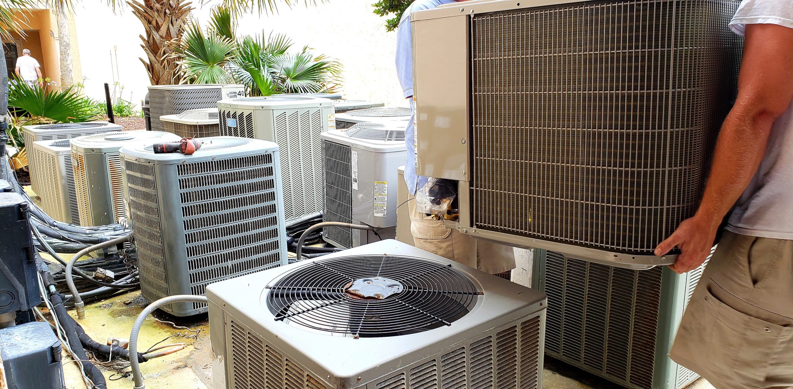 Technician standing among multiple outdoor air conditioning units in a sunny courtyard.