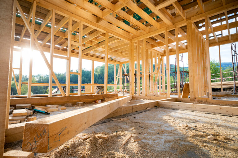Interior of a wooden-framed building under construction with exposed studs and beams, sunlight streaming in through gaps.