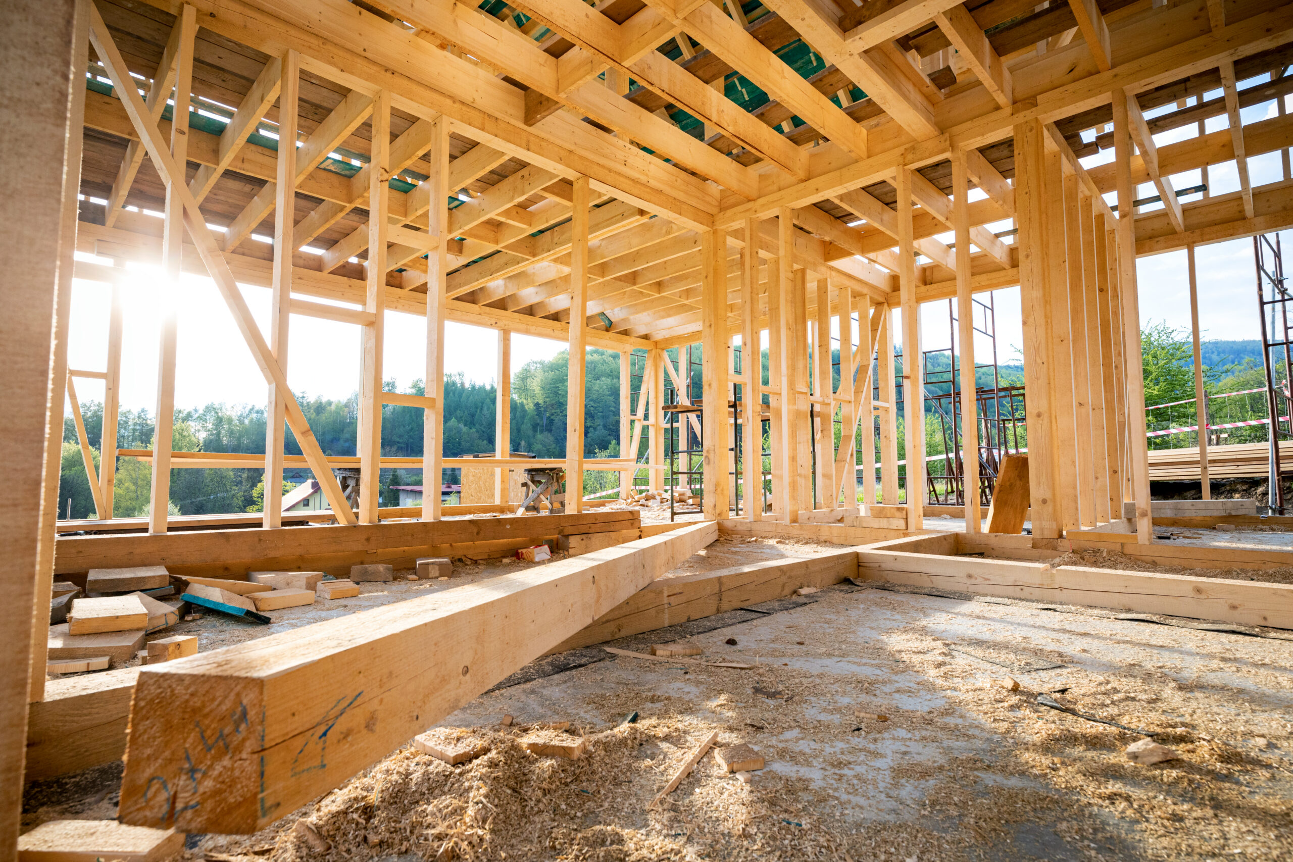 Interior of a wooden-framed building under construction with exposed studs and beams, sunlight streaming in through gaps.