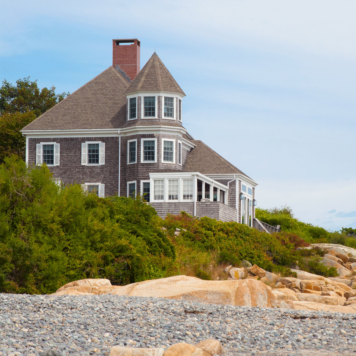 Coastal wooden house with a turret perched on a rocky beach, surrounded by green shrubs under a blue sky as waves nearby.