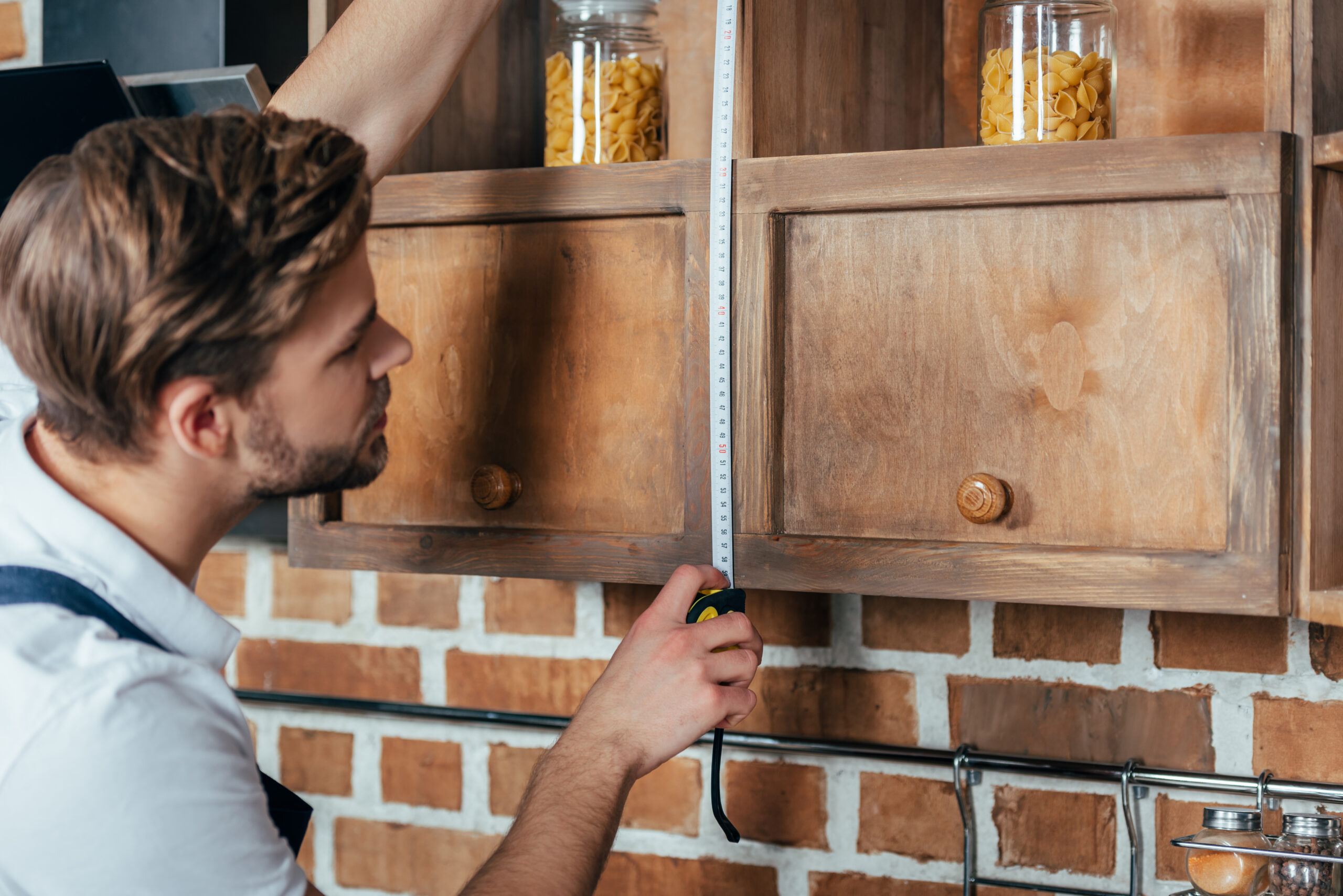 Man measuring the height of a wooden cabinet with a tape measure in a workshop setting.