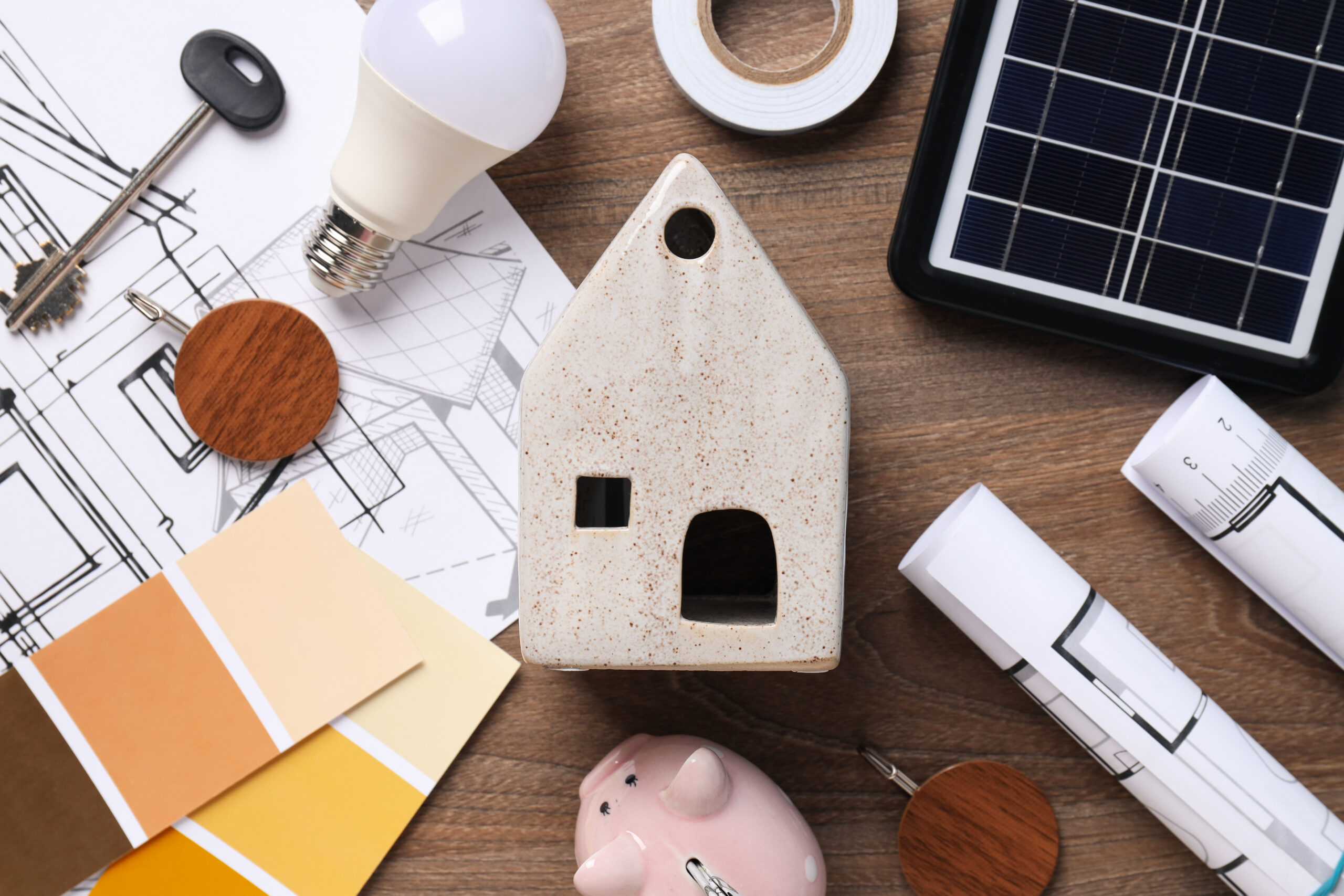 Architectural workspace with blueprints, a ceramic house model, a light bulb, a solar panel, and color swatches on a wooden desk.